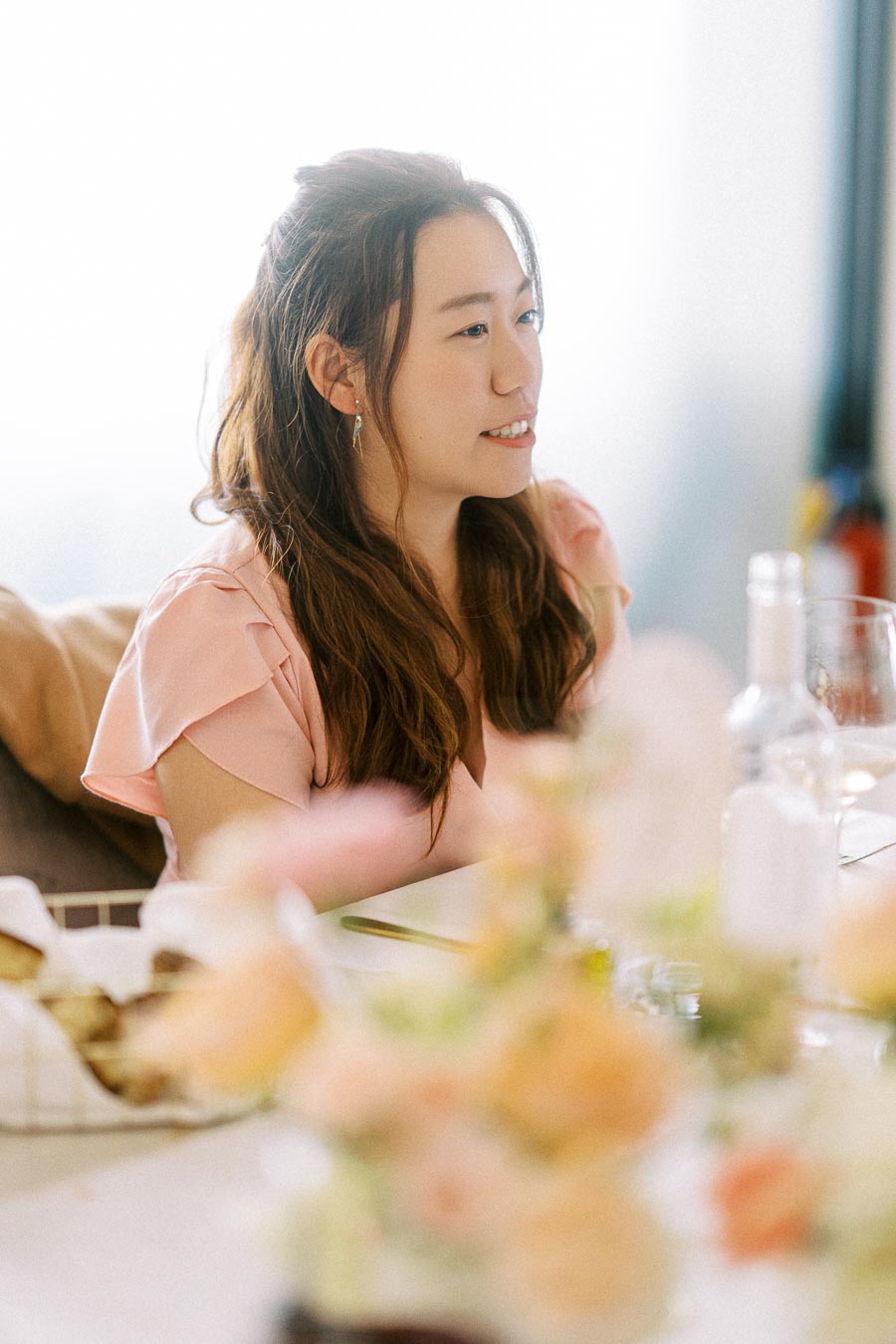 Young woman with long hair sitting at a dining table, smiling and wearing a light pink blouse, surrounded by blurred flowers