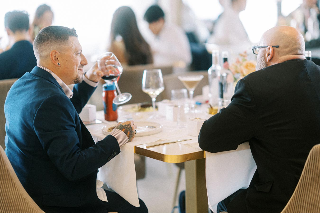 Two men in suits enjoying a conversation over drinks at a restaurant table, with wine and cocktails visible; a casual dining