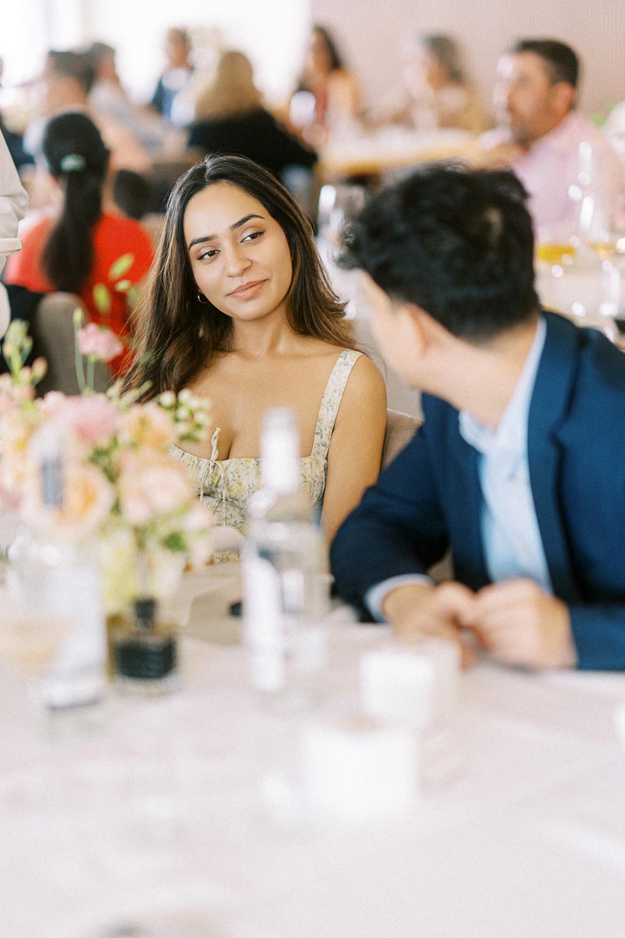 Two people engaging in a conversation at a social event, surrounded by a blurred audience. A beautifully arranged bouquet of