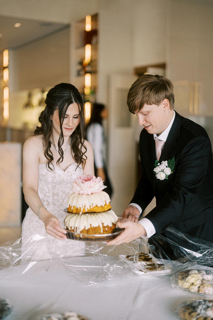 Wedding couple placing a tiered bundt cake on a table with decorative icing, surrounded by dessert platters in an elegant