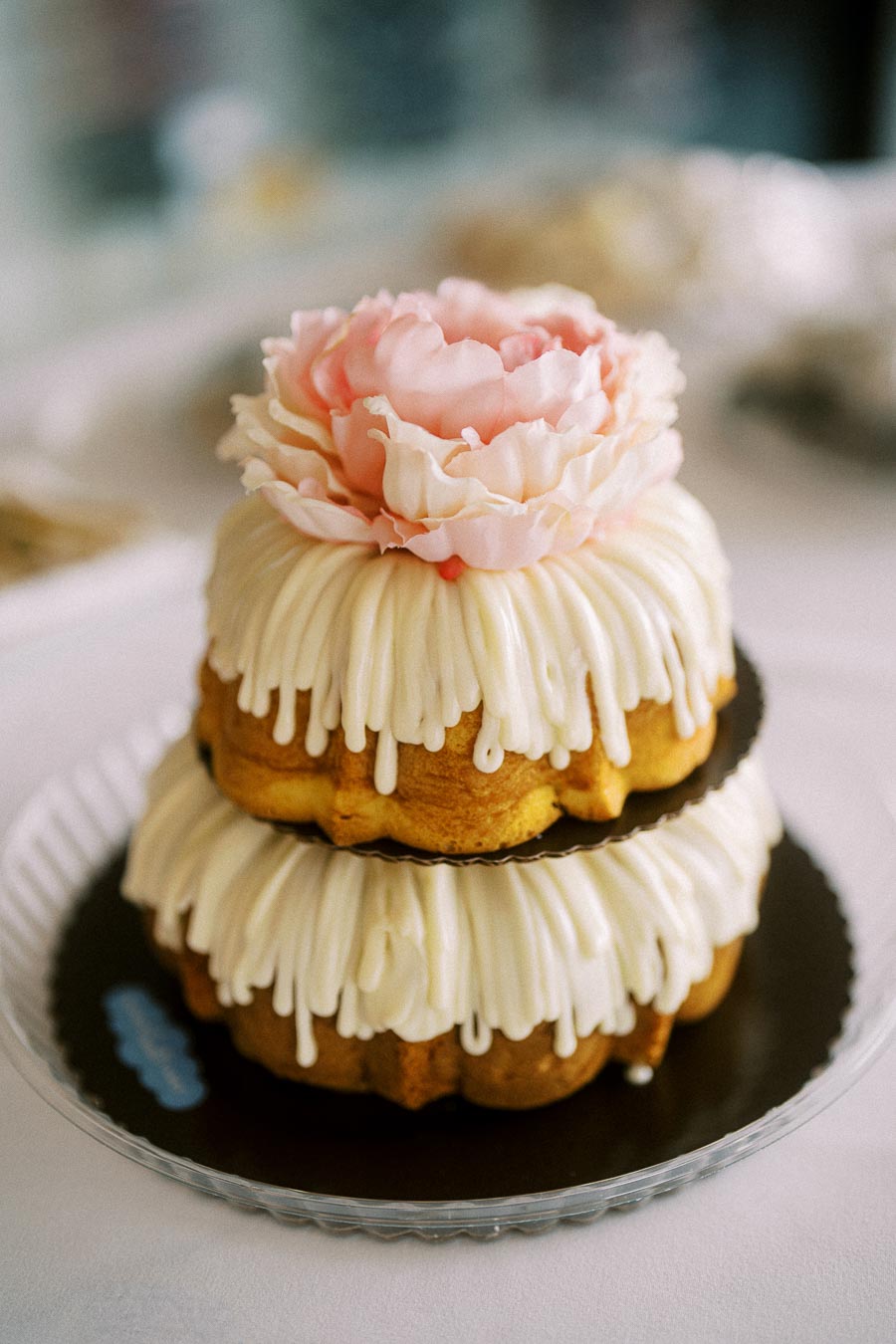 Delicate two-tier bundt cake with creamy white icing, adorned with a pink flower on top, elegantly displayed on a clear