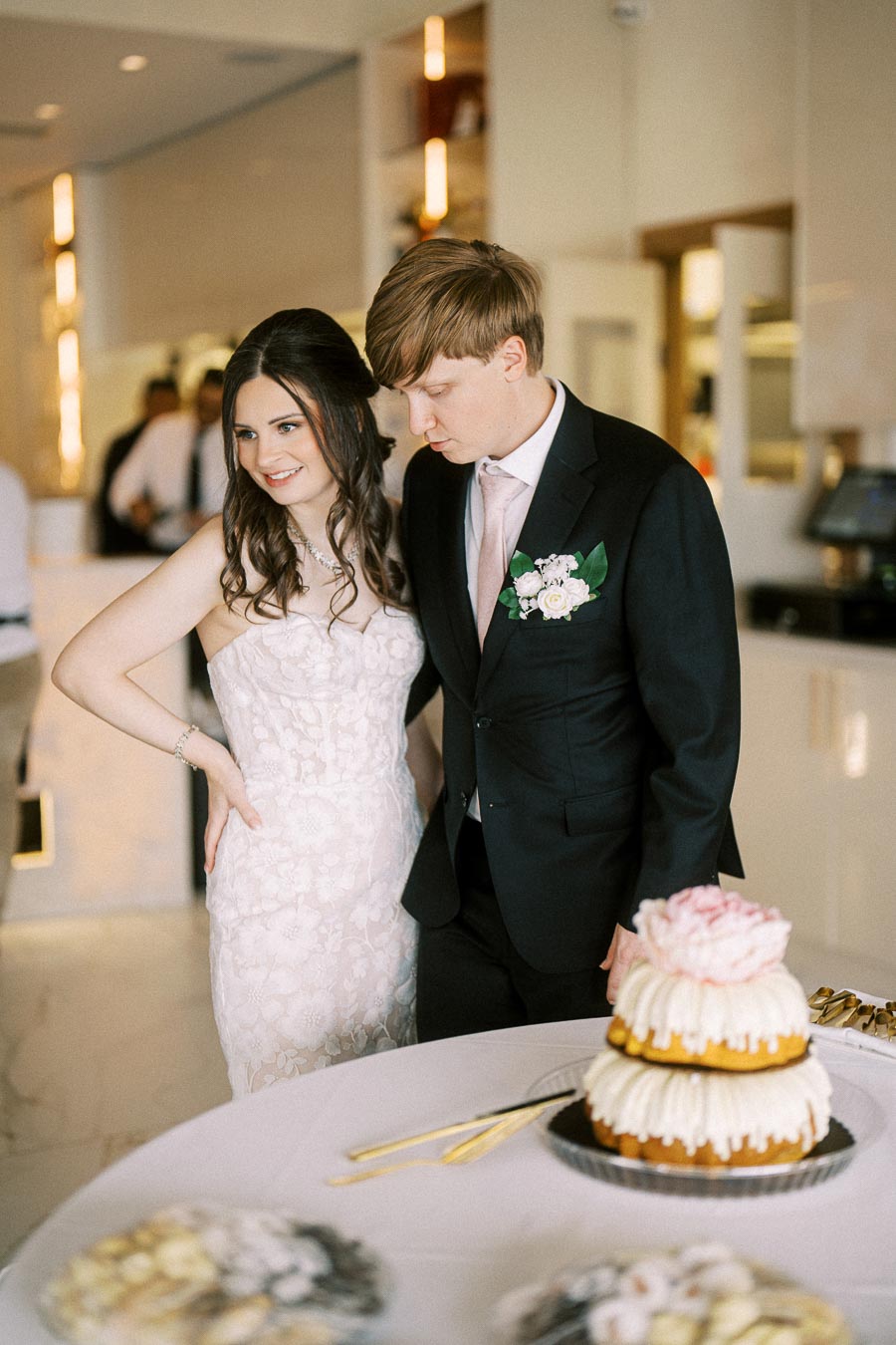 Bride and groom smiling at a tiered wedding cake with floral decoration, indoors at a wedding reception.