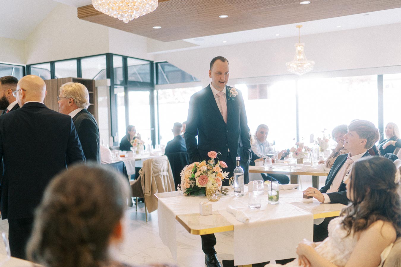 Wedding reception scene in a bright, elegant venue with guests in formal attire. Central figure standing at a table adorned