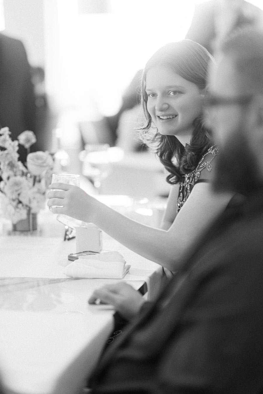 Black and white image of a woman smiling while seated at a table during a social event, holding a glass with floral