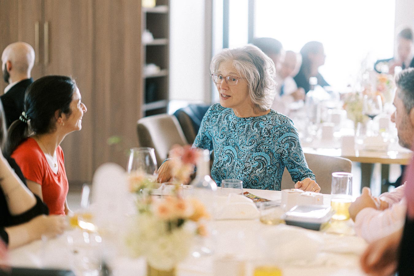 A group of people engaged in a lively conversation around a dining table, featuring a woman in a blue patterned blouse