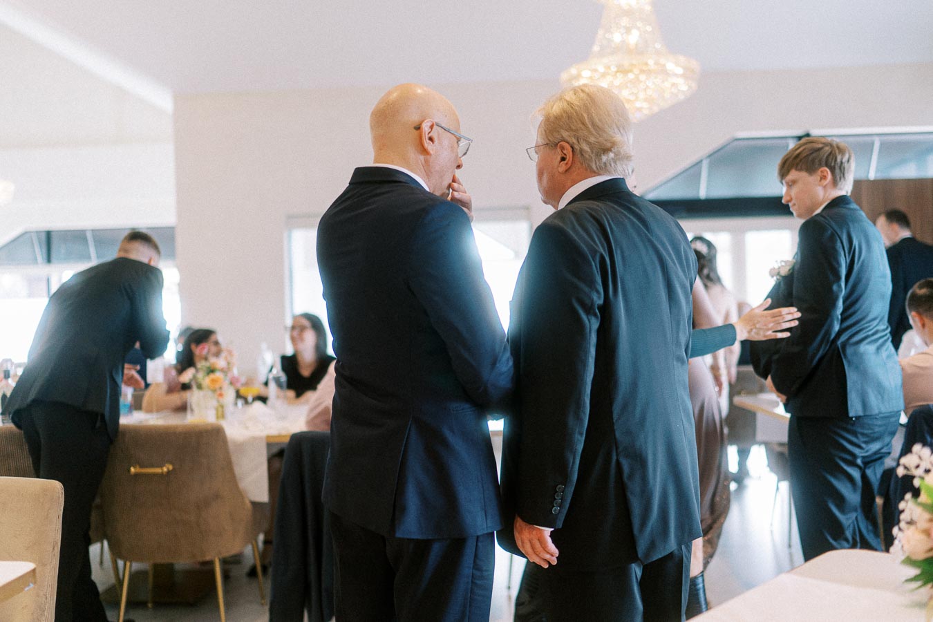 Group of people in formal attire engaging in conversation at an indoor event, with tables set up for dining in the