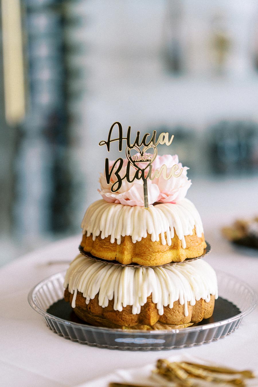 Two-tiered bundt cake with white icing, topped with a custom gold cake topper, displayed on a clear plate.