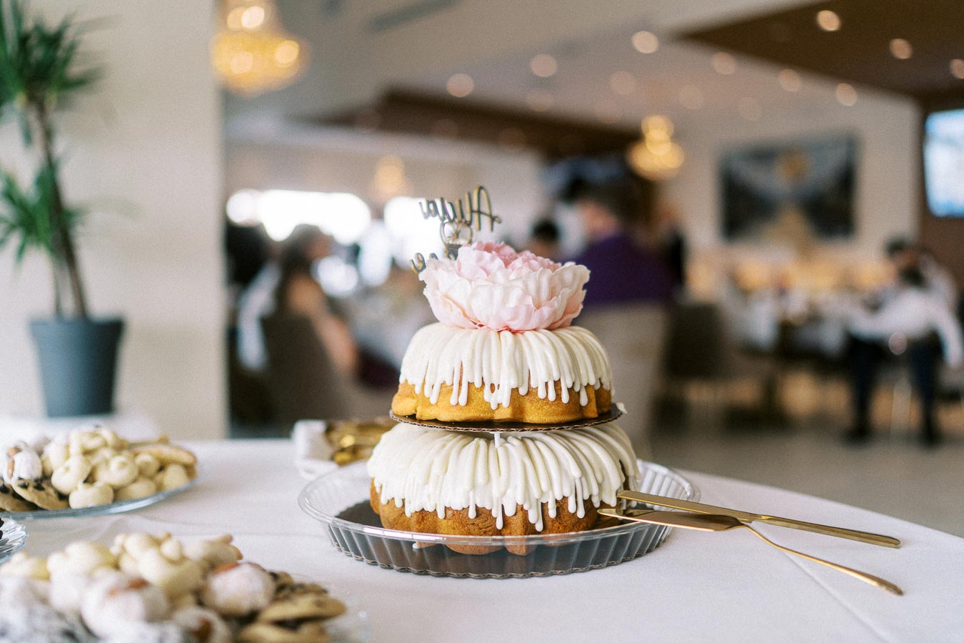 A two-tiered bundt cake with white icing and a pink flower decoration on top, displayed in a bright, elegant dining setting,