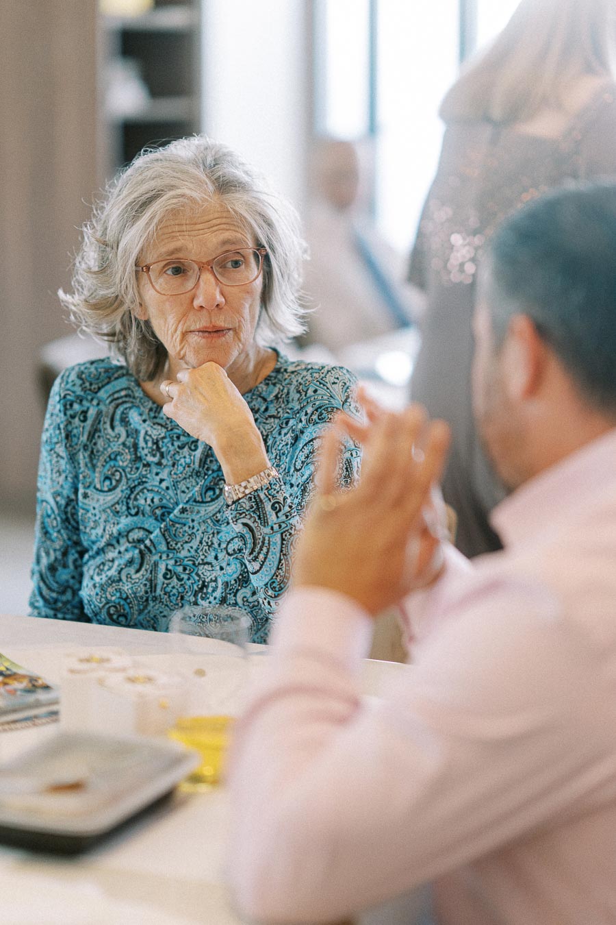 Elderly woman in a blue patterned blouse in conversation with a man at a gathering, sitting at a table with blurred