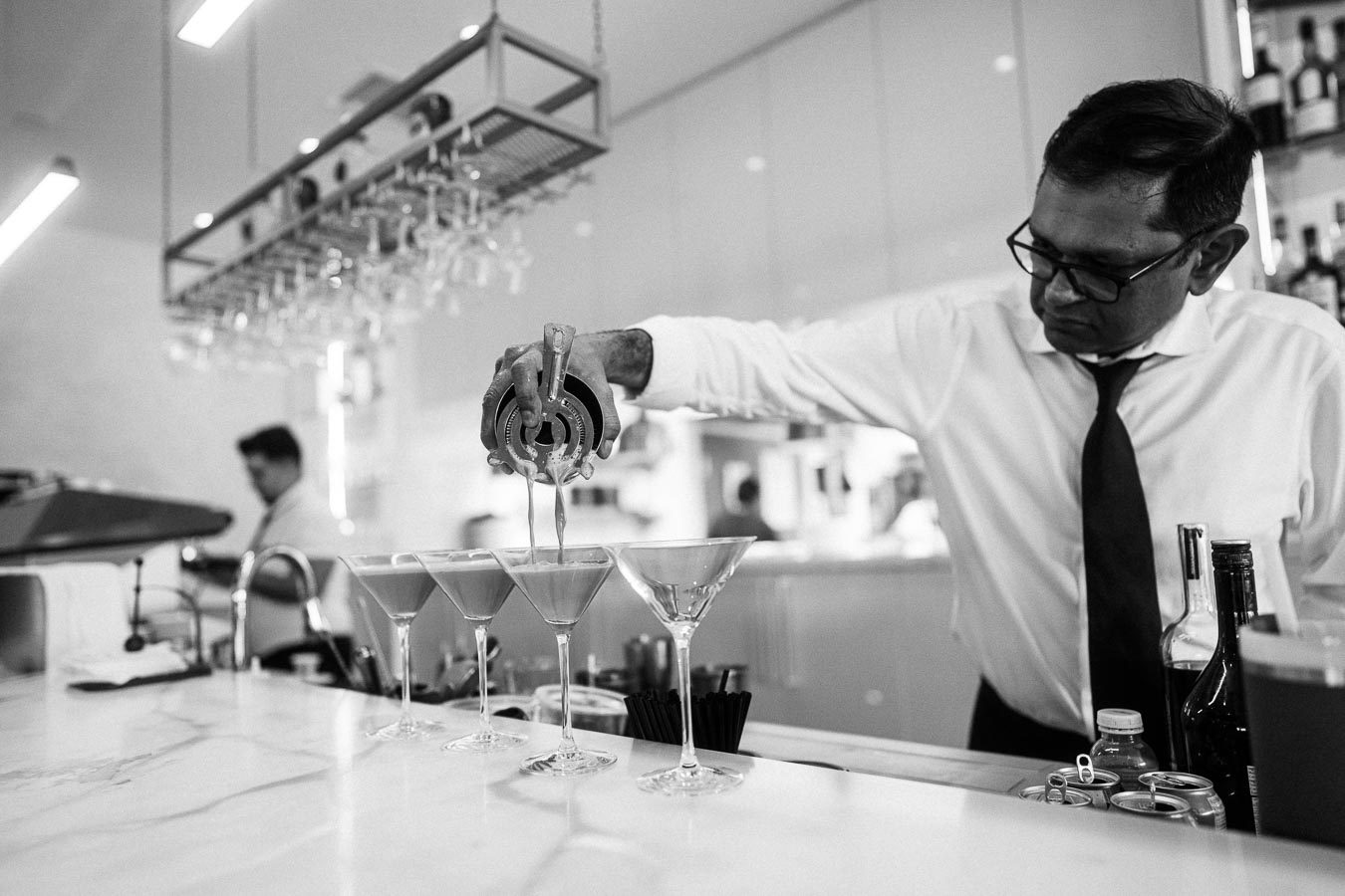 A bartender in a white shirt and tie skillfully pours a cocktail into glasses at a modern bar, with bottles and bar tools