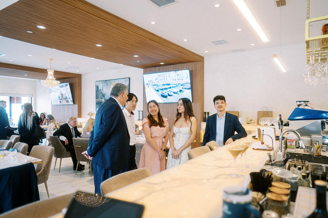 Group of people socializing in a modern restaurant setting, featuring a bar counter with various drinks and kitchenware,