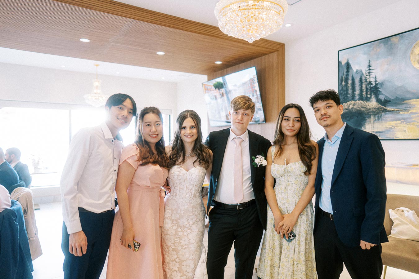 A group of six elegantly dressed friends posing together at a wedding reception, with a bride in a white lace gown and a