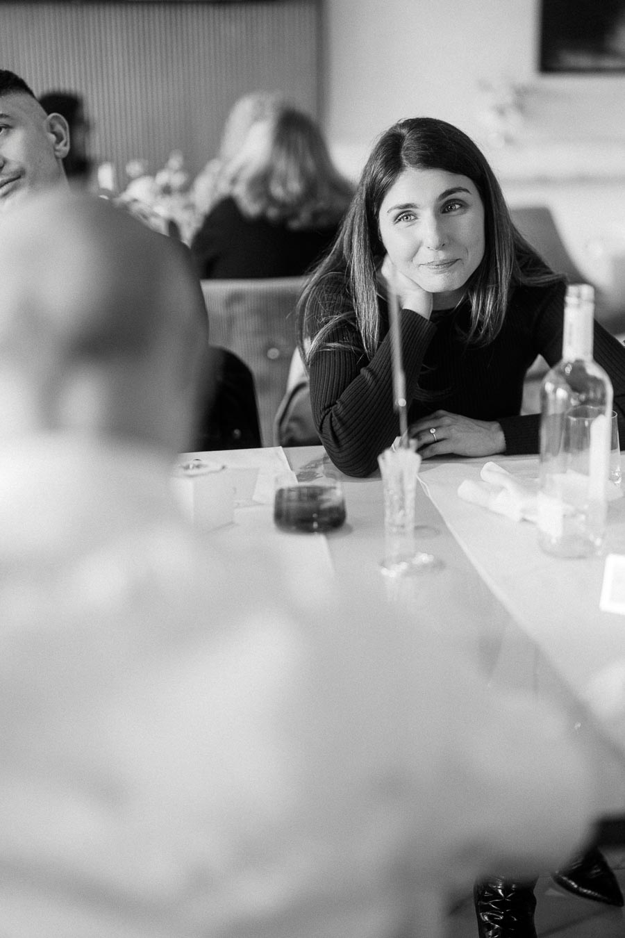 A woman with long hair sits at a table, looking thoughtfully across at someone. The setting appears to be a social