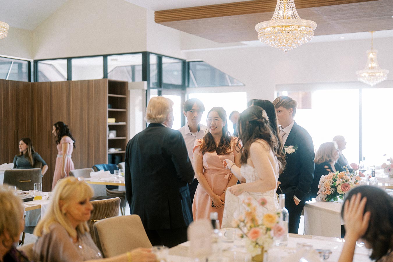 Group of people celebrating at an indoor event, dressed in formal attire, with decorative chandeliers and floral