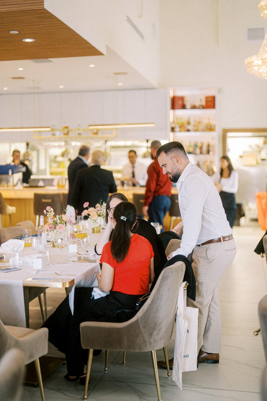 People socializing in a modern restaurant with elegant decor, flowers on the table, and staff in the background.