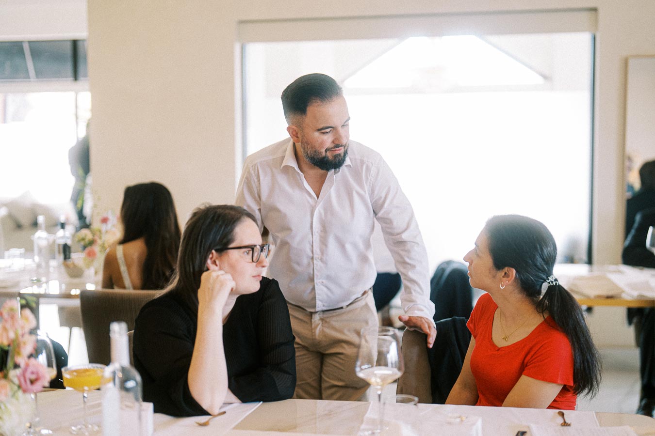 A group of people engaged in conversation at a restaurant table with drinks and floral decorations, featuring a relaxed and