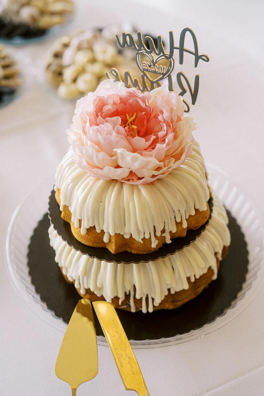 Two-tier bundt cake with white icing, adorned with a large pink flower topper and elegant gold utensils, displayed at an