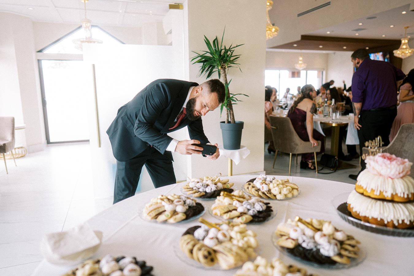 Man in a suit photographing a dessert table at a formal event, featuring a variety of pastries and a tiered cake, with