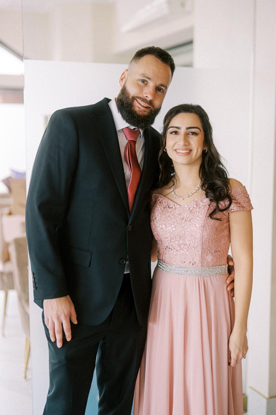 A smiling couple dressed in formal attire, with the man wearing a dark suit and red tie, and the woman in a pink lace gown,