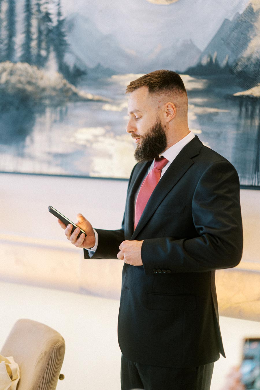 A man in a formal suit with a red tie looking at a smartphone in an artfully decorated room.