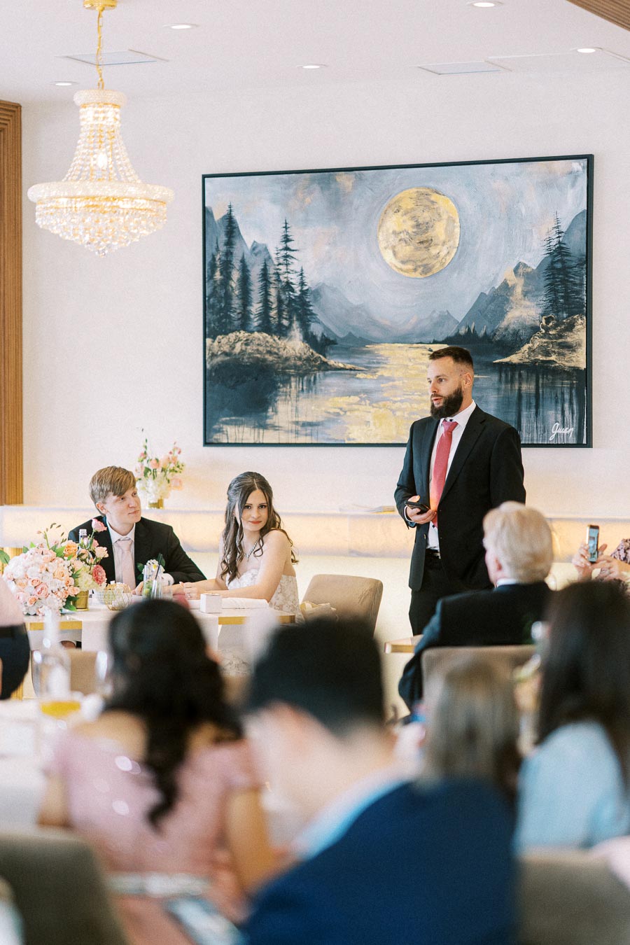 Wedding reception speech with guests seated elegantly at decorated tables, featuring a prominent landscape painting on the