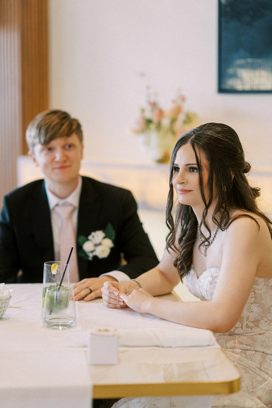 A couple sitting at a table, both dressed in formal wedding attire. The woman is wearing a white lace bridal gown and the