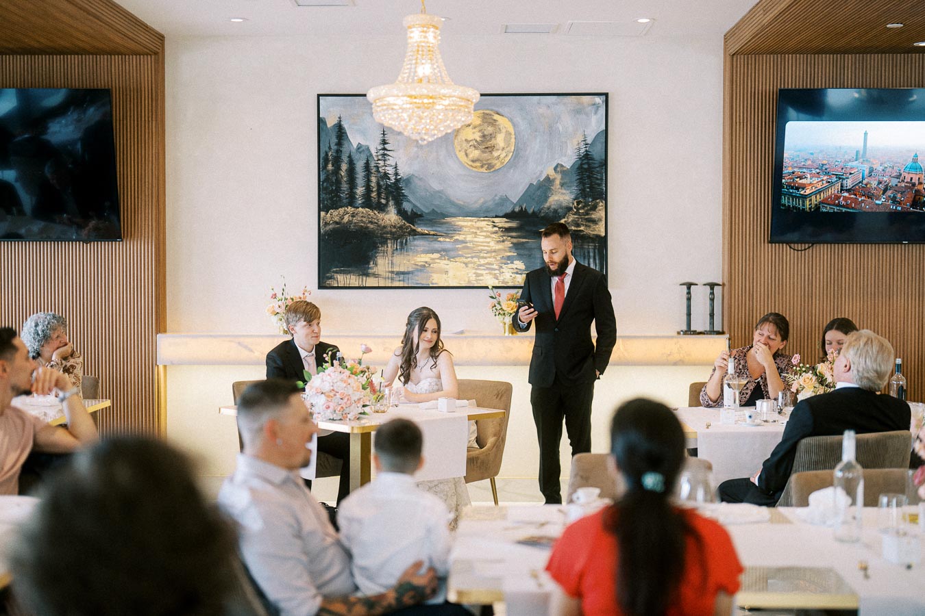 A man in a suit gives a speech at a wedding reception in an elegantly decorated room, with guests seated at tables and a