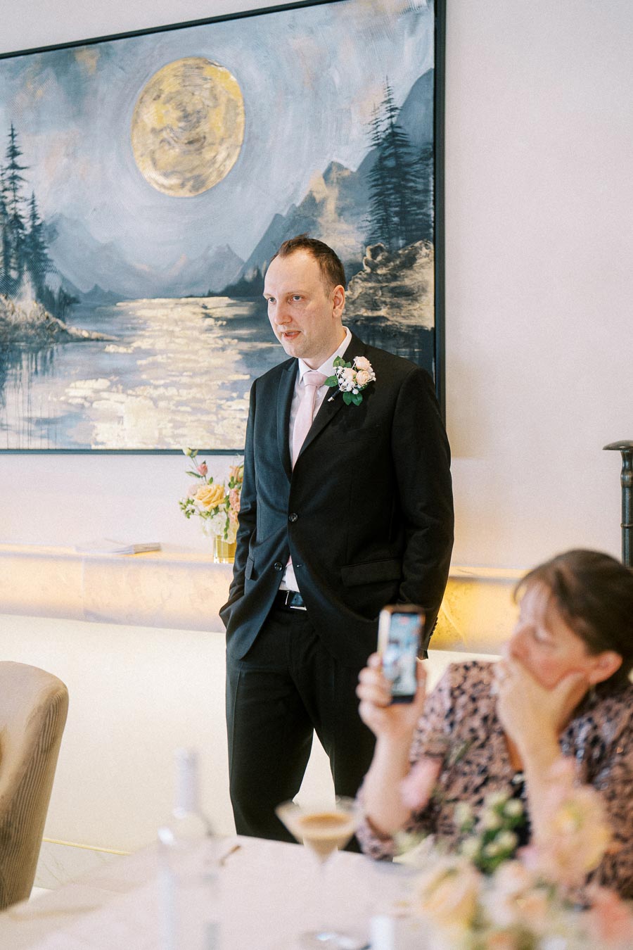 Man in a suit with pink tie and boutonniere standing in front of a large painting, with a woman in the foreground taking a