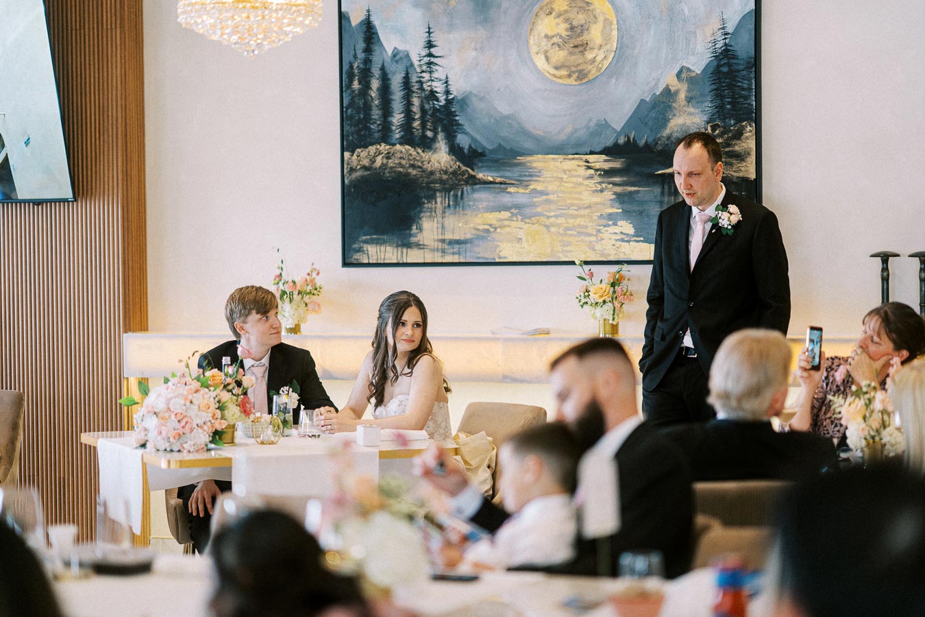 Wedding reception scene with a sitting couple and a standing man, decorated with floral arrangements and a large painting of