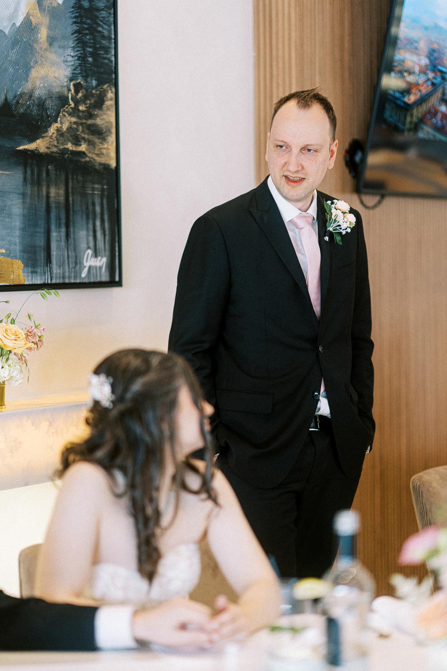 A man in a suit giving a speech at a wedding reception, with seated guests listening attentively. Elegant decor and floral