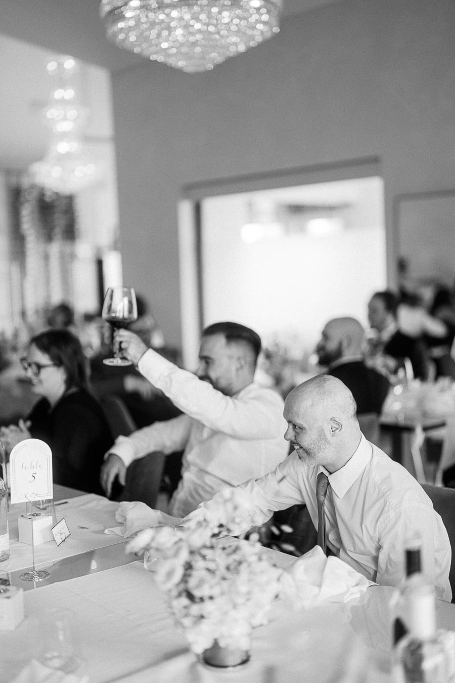 Black and white photo of guests at a formal event. A man in a white shirt raises a wine glass in celebration, seated at an