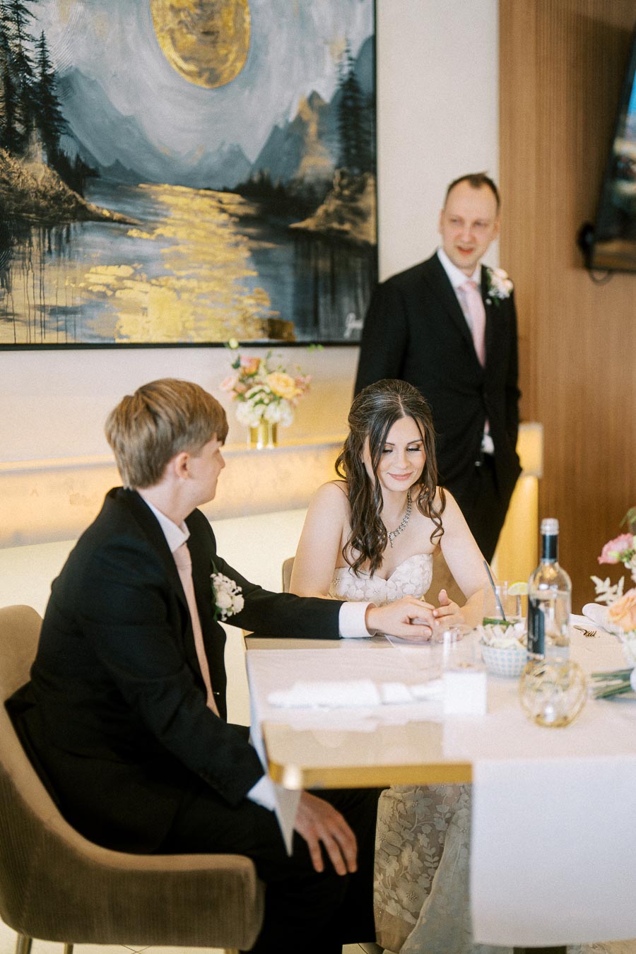 A bride and groom seated at a wedding reception, smiling and holding hands, with a decorative painting and flower