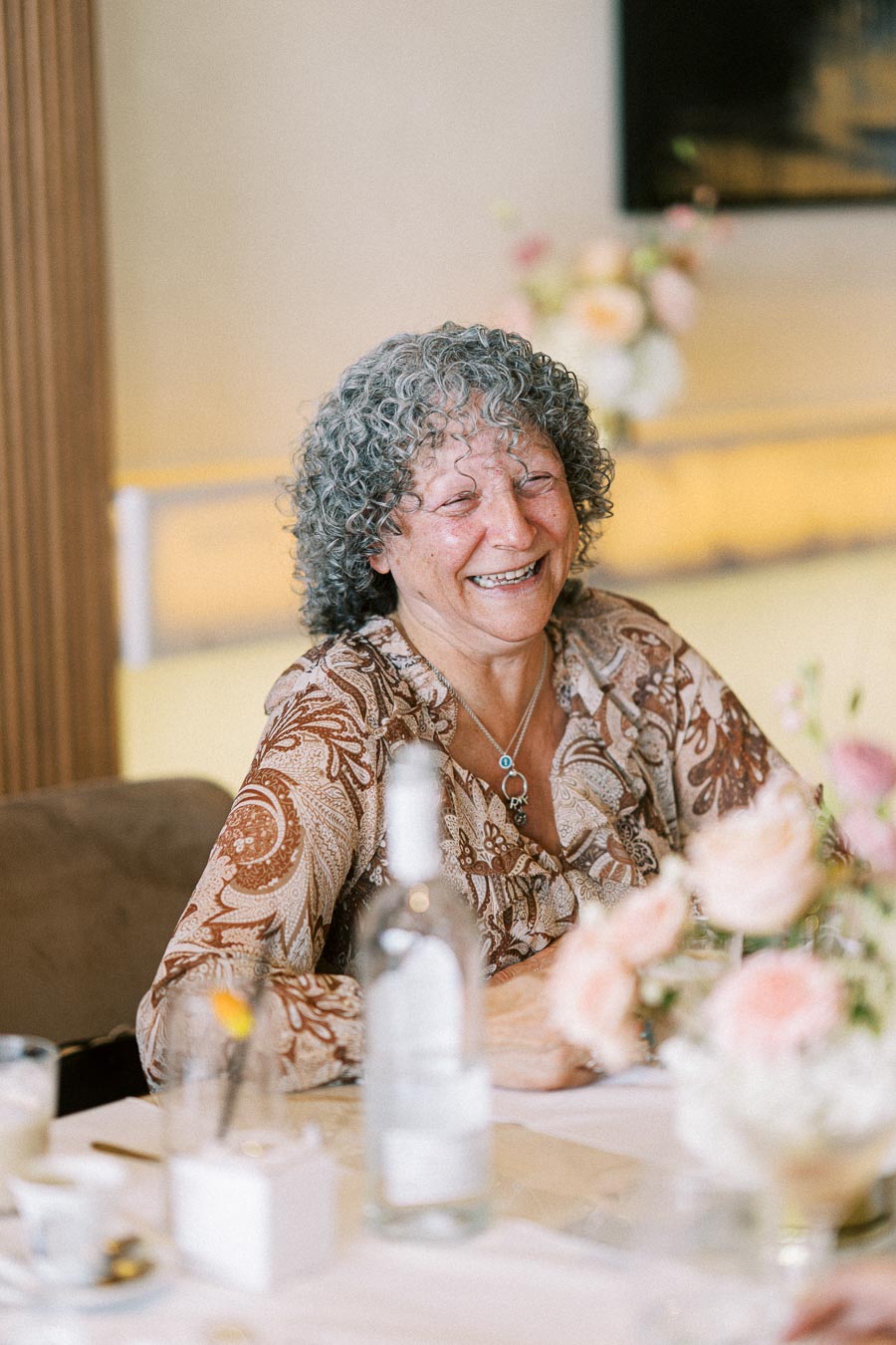 Elderly woman smiling at a beautifully set table, wearing a patterned blouse and necklace, surrounded by soft pastel flowers