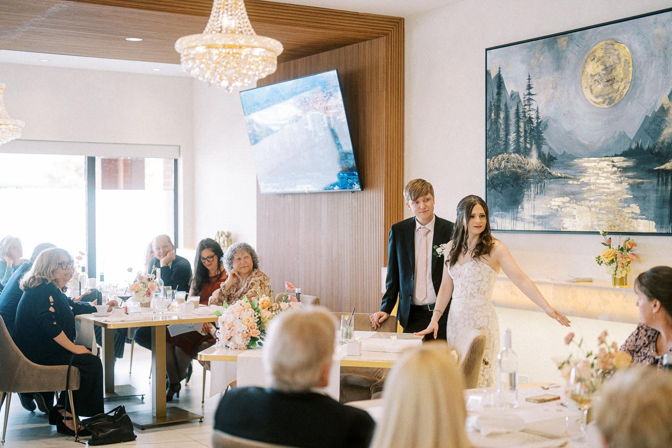 A bride and groom addressing guests at a wedding reception in an elegantly decorated room with chandeliers and a large