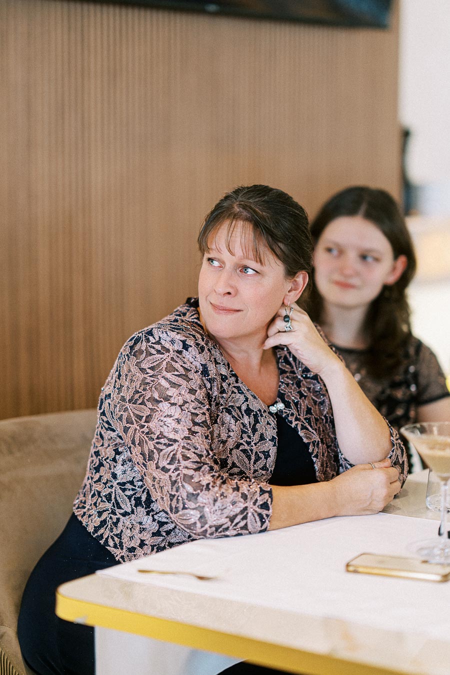 Two women seated at a table, one in a floral lace jacket thoughtfully looking away, with a cocktail glass and mobile phone