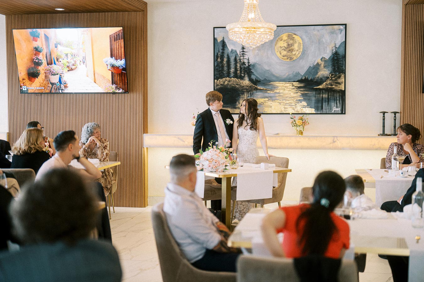 A couple standing at the head table during a wedding reception, surrounded by guests in a beautifully decorated venue with