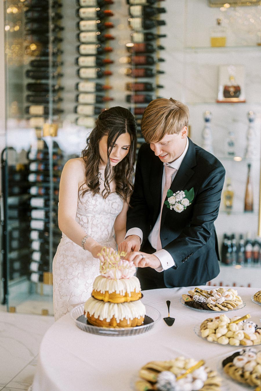 A couple cutting a tiered wedding cake adorned with icing and flower decoration, surrounded by a selection of pastries and