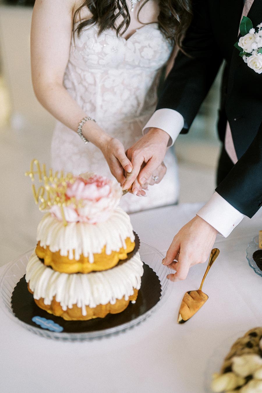 A bride and groom cutting a two-tiered wedding cake with white icing and floral decoration, using a gold cake knife during