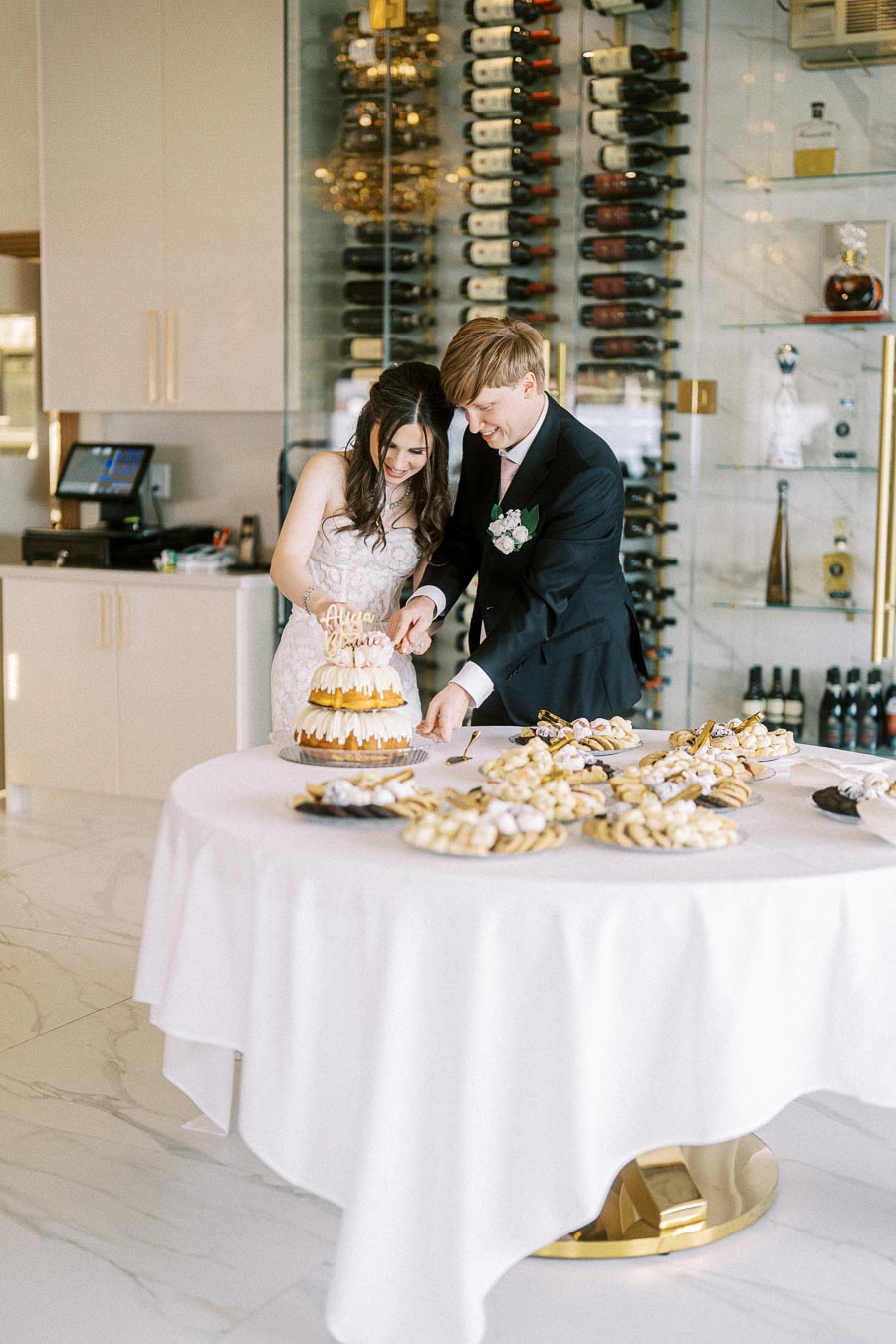A bride and groom cutting a tiered wedding cake at a reception with a dessert table, surrounded by wine bottles in the