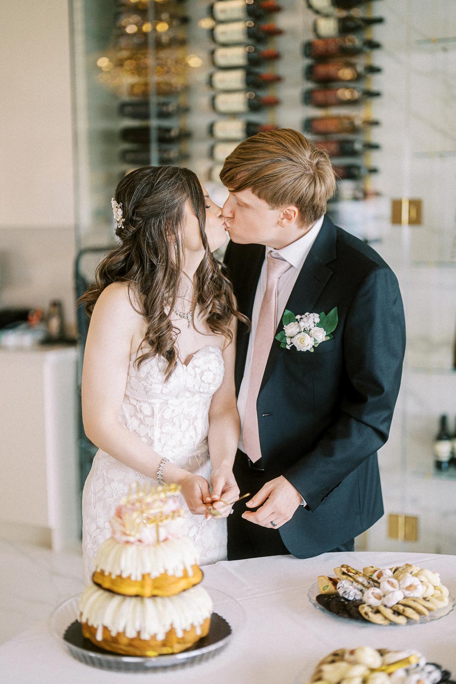 A couple in wedding attire shares a kiss while cutting a tiered cake, surrounded by a wine display and assorted desserts.