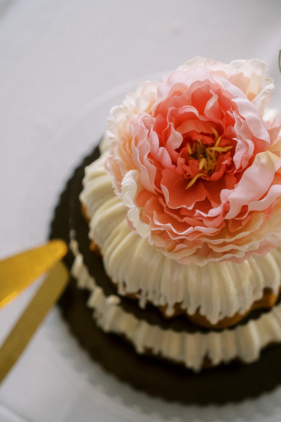 A beautifully decorated cake featuring a large pink peony flower on top, with intricate white icing details, resting on a