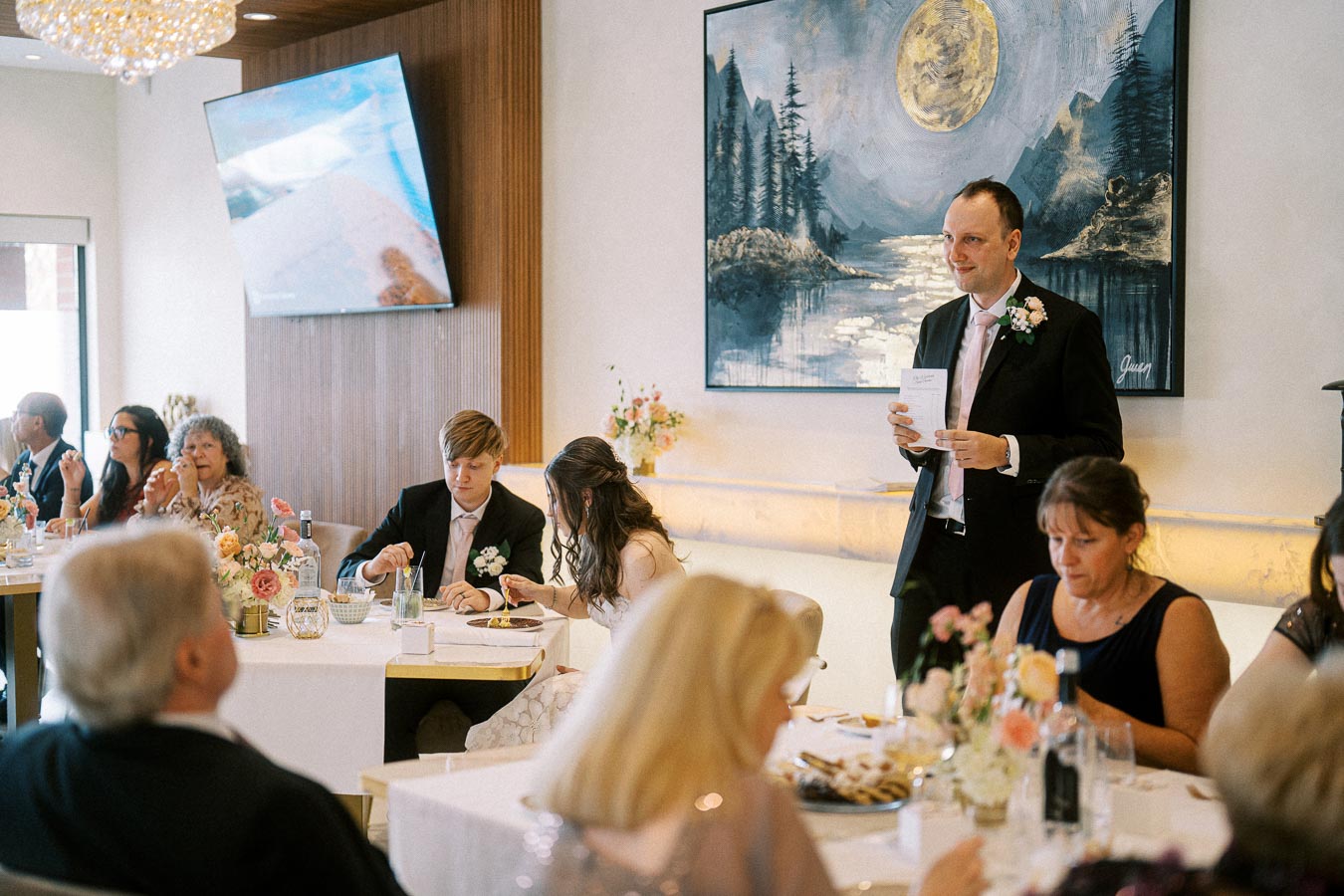 Wedding reception scene in a decorated venue, featuring a man giving a speech while guests are seated at elegantly set