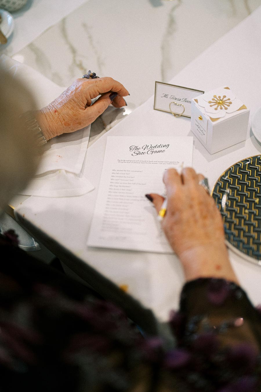Elderly woman participating in a wedding shoe game, reading instructions at a decorated table with a name card and gift box.