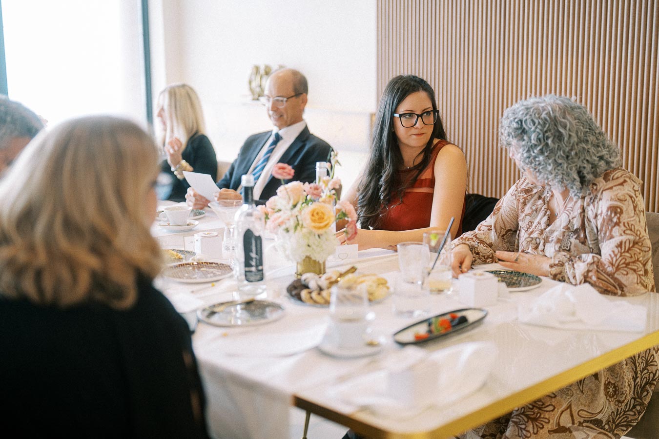 People gathered around a dining table at a formal event, with elegant tableware and a floral centerpiece.