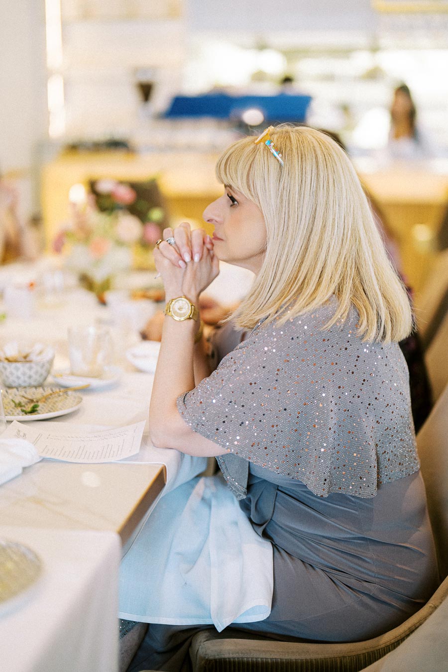 A woman with blonde hair sits at a dining table, wearing a sparkling grey shawl and gold watch, gazing thoughtfully to the