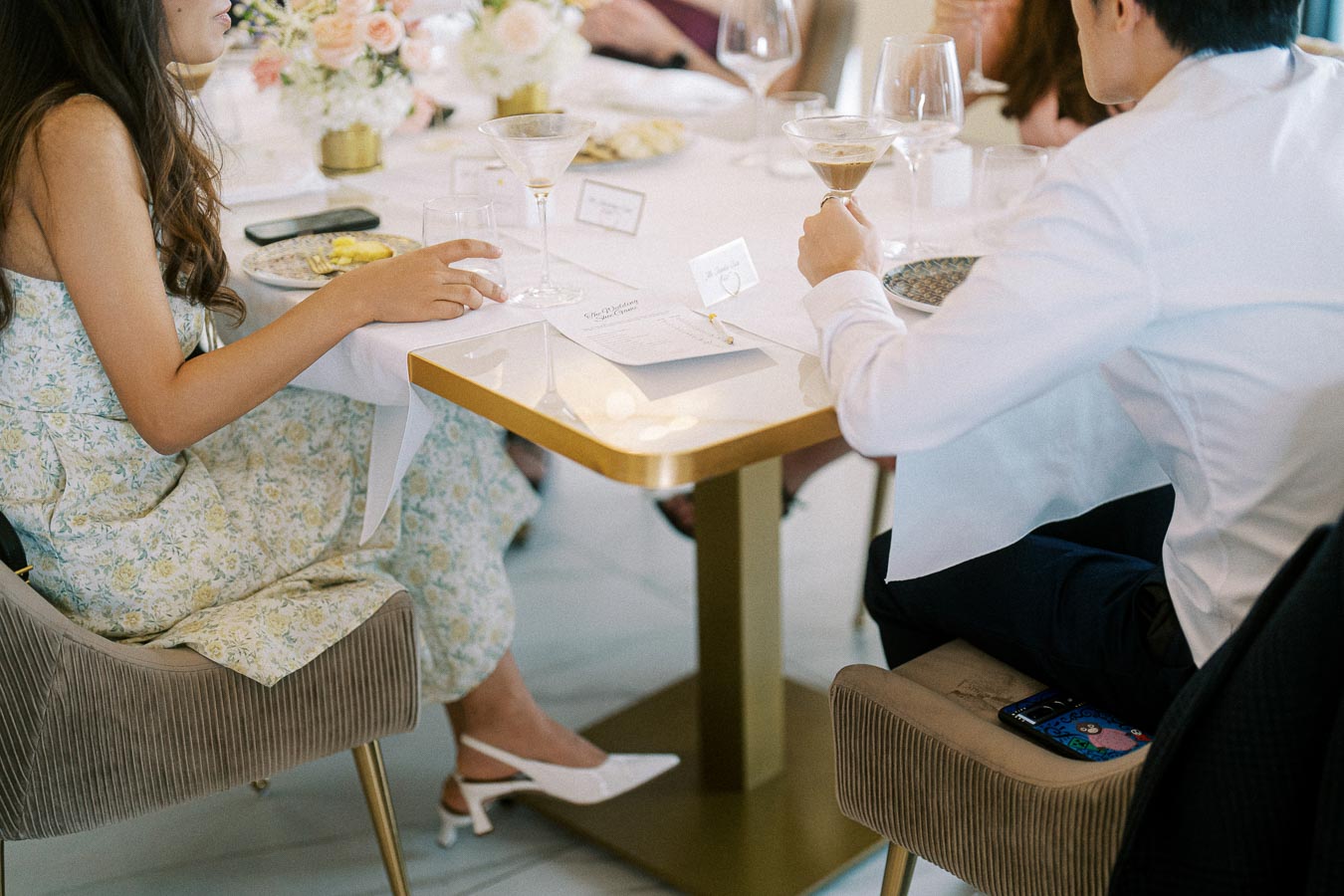 Two individuals seated at an elegant dining table, enjoying a sophisticated meal with drinks, surrounded by floral