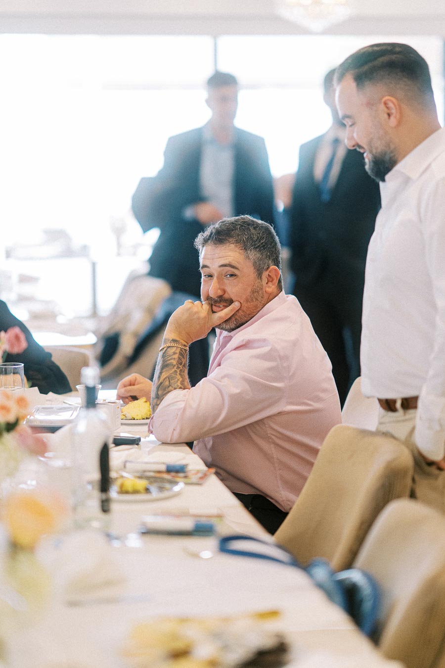 A man in a pink shirt with a tattoo on his arm smiling and seated at a dining table during a gathering or event, surrounded