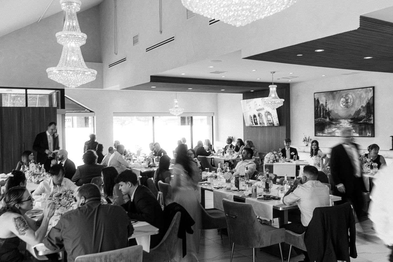 Black and white image of a formal gathering in a elegantly decorated banquet hall. Guests are seated at tables adorned with