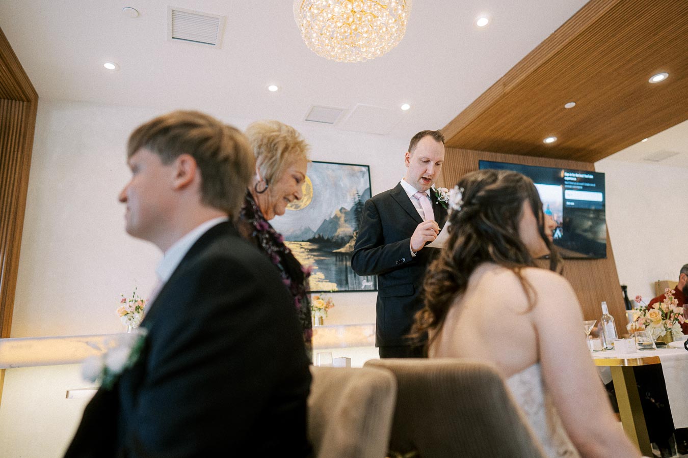 A man in a suit gives a speech at a wedding reception, surrounded by guests seated at tables with floral decorations, under