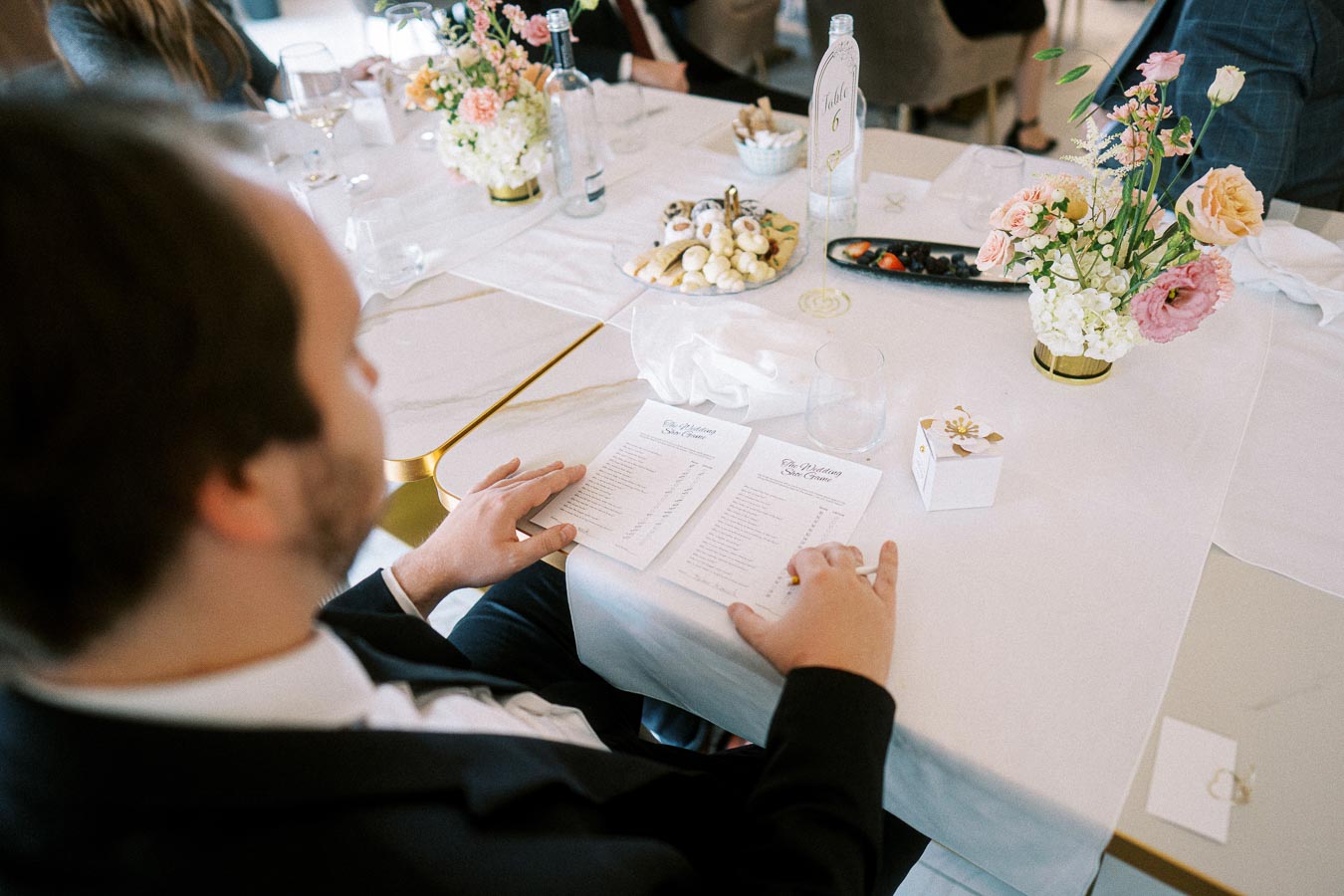 Man in formal attire reviewing menu at a wedding table setting with elegant floral centerpiece, decorative tableware, and